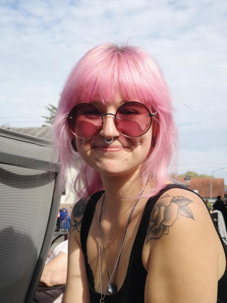 Person with pink hair and rose-tinted sunglasses smiling outdoors